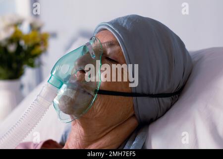 Senior caucasian female cancer patient wearing head scarf in bed with oxygen mask at hospital Stock Photo