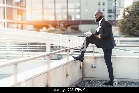 View of a fancy mature bald black businessman in a tailored formal suit and eyeglasses putting one foot on the ledge so that it would be convenient to Stock Photo