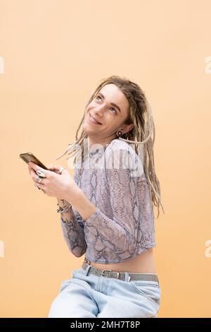 Smiling queer person in crop top with snakeskin print and jeans using smartphone isolated on yellow,stock image Stock Photo