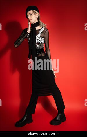 full length of smiling nonbinary person in black elegant clothes holding hand near face on red background with shadow,stock image Stock Photo
