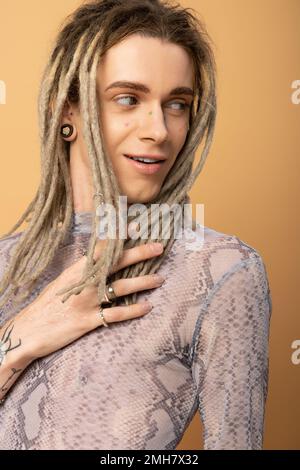 smiling queer person in top with snakeskin print touching dreadlock and looking away isolated on yellow,stock image Stock Photo