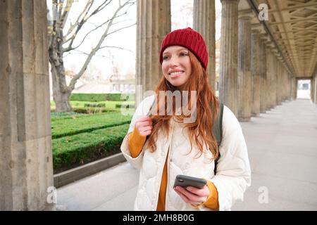 Mobile broadband and people. Smiling redhead 20s girl with backpack, uses smartphone on street, holds mobile phone and looks at application Stock Photo