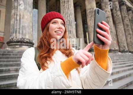 Portrait of young blogger, redhead girl tourist takes pictures of sightseeing, makes photos on smartphone camera, records video and smiles Stock Photo