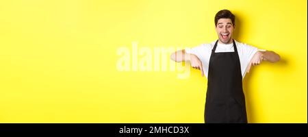 Excited waiter in black apron pointing fingers down, checking out promo offer, standing over yellow background Stock Photo