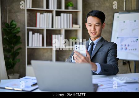 Handsome and happy millennial Asian businessman or male financial consultant using his smartphone at his desk. Stock Photo