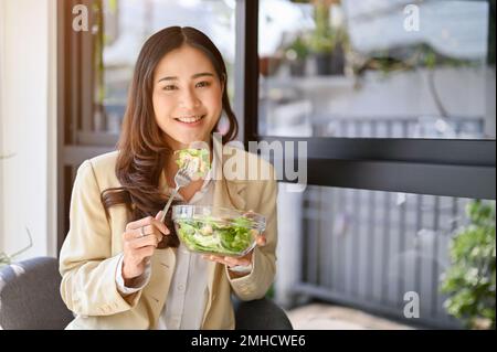 Beautiful millennial Asian businesswoman having brunch, holding a salad bowl, enjoying eating healthy salad bowl at the cafe. Stock Photo