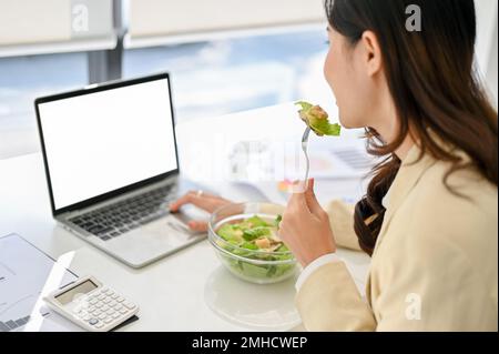 close-up image of a busy millennial Asian businesswoman eating healthy salad bowl, having lunch while working on her project on laptop in the office. Stock Photo