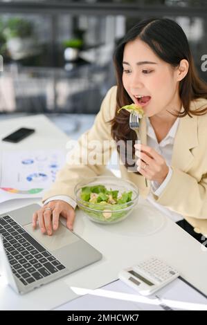 Portrait, Hungry and busy millennial Asian businesswoman eating salad bowl, having lunch while working on her business tasks on laptop. overwork, mult Stock Photo