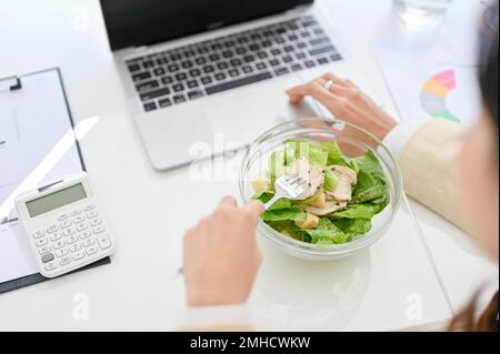 close-up image of a busy millennial Asian businesswoman eating healthy salad bowl, having lunch at her desk while managing her business tasks on lapto Stock Photo