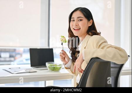 Charming millennial Asian businesswoman is at her desk with a healthy salad bowl, having lunch at her office desk. Stock Photo