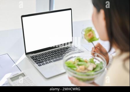close-up image of a busy Asian businesswoman eating healthy salad bowl, having lunch at her desk while managing her business tasks on laptop. laptop w Stock Photo
