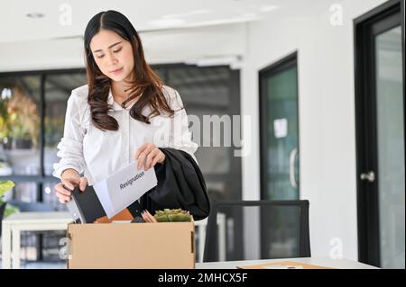 Sad and depressed young Asian female office worker packing her stuff into a cardboard box in the office, quitting her job or getting fired from her bo Stock Photo