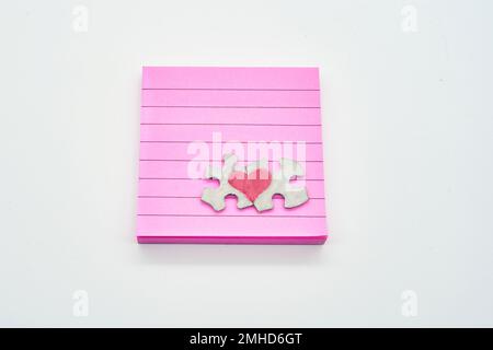 Heart on connected puzzle pieces on top of post it note pink note pad on solid white background Stock Photo