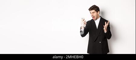 Portrait of angry businessman in black suit yelling at mobile phone, having an argument on video call, standing mad over white background Stock Photo