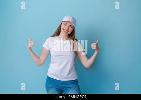 Hey look. Smiling beautiful female model pointing her fingers to an empty space and inviting to check it out showing an advertisement on a blue background. Dressed in white t-shirt and cap for mockup Stock Photo