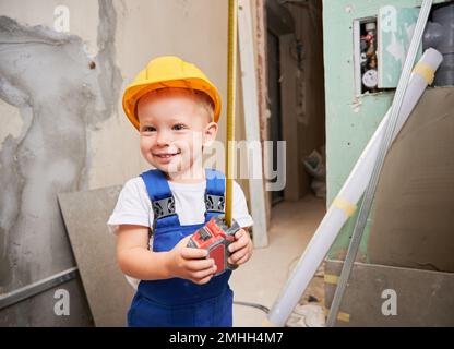 Close up of boy construction worker holding measuring tape and smiling while standing in apartment under renovation. Cheerful kid with measuring tool wearing safety helmet and work overalls. Stock Photo