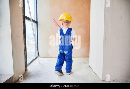 Child checking wall surface with spirit level in apartment. Back view of kid in work overalls and safety construction helmet using level instrument while working on home renovation. Stock Photo
