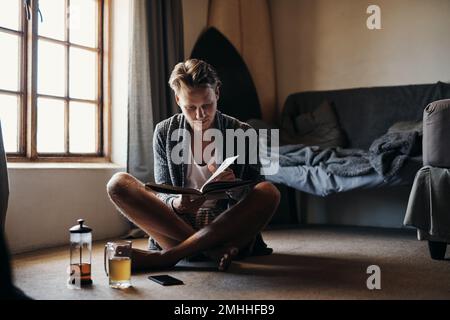 This book is truly amazing. a young man reading a book while sitting on the floor at home. Stock Photo