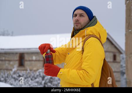 Thoughfull man in a yellow jacket holds thermos with hot drink in hands on snow winter day outdoors. Stock Photo
