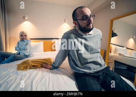 Worried man sit on bed in bedroom, his angry wife looks at him. Stock Photo