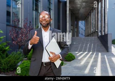 Portrait of successful african american man outside office building, man in suit and glasses standing and looking at camera smiling and giving affirmative thumbs up. Stock Photo