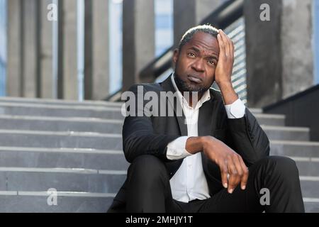 Upset african american man sitting on stairs outside office building, businessman bankrupt in despair, mature boss depressed in business suit outdoors. Stock Photo