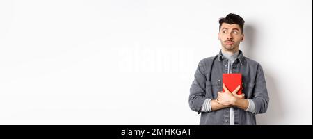Silly young man looking aside at logo with timid face, holding his diary pressed to chest, carry journal, standing on white background Stock Photo