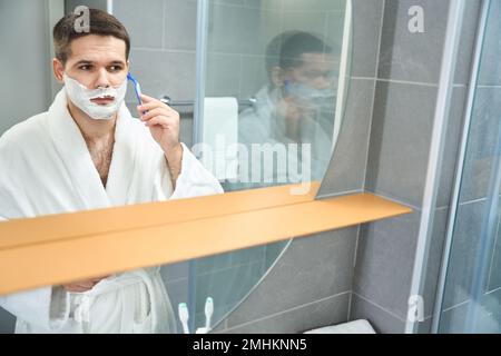 Man in a bathrobe shaves his cheek with a razor Stock Photo