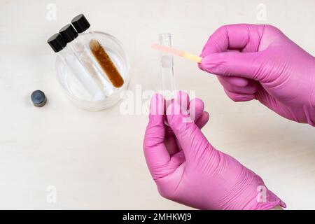 gloved hands holding test tube with liquid and indicator paper showing pink color, acid pH level over light desk Stock Photo