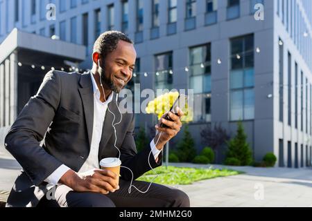 African american businessman using phone sitting on bench outside office building, boss watching video and smiling in headphones, man outdoors listening music audiobook and podcasts online radio. Stock Photo