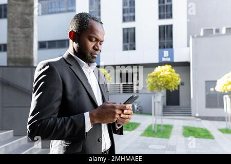 Serious and thoughtful businessman reading online news on phone african american boss in suit outside office building looking at smartphone screen concentration, walking near building in background. Stock Photo