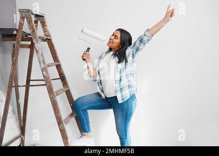 Wide shot of young Indian woman singing with paint roller while painting wall Stock Photo