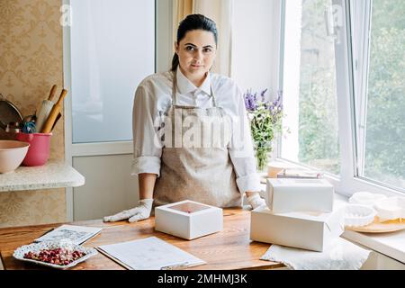 Online classes and workshops on cake decorating and baking. Female Confectioner baker make custom pink heart shape cake in kitchen Bakery. Stock Photo