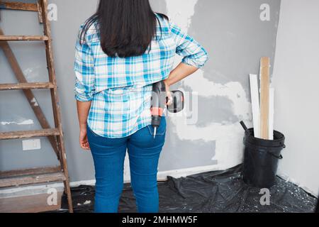 Cropped shot of back of a woman holding drill on her hips while looking at wall Stock Photo