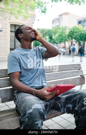 Man smiling while using a digital tablet sitting on a bench on the street. Stock Photo