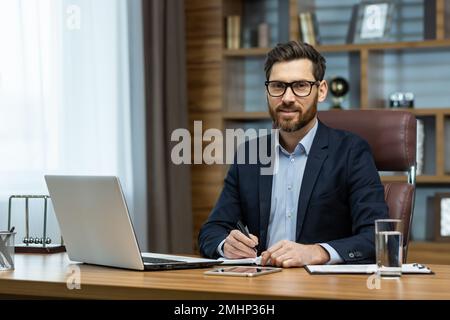 Portrait of successful smiling man in office, mature businessman looking at camera cheerfully, senior boss in glasses and beard working inside office with laptop and documents, signing contract. Stock Photo