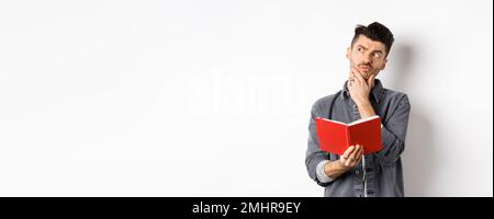 Thoughtful young man plan his schedule, looking aside pensive, holding opened planner or journal, standing on white background Stock Photo
