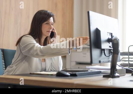 Executive female manager in a suit works at a computer in the office and gives tasks to her team. Stock Photo