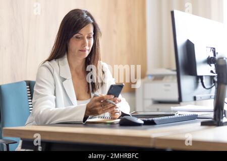 A manager in a suit works at a computer in the office and reads the messages on a mobile phone. Stock Photo