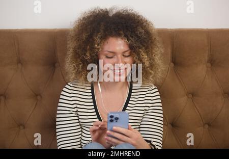 Happy young woman enjoying music in headphones. Cheerful curly female person sitting in bedroom and listening to favorite musical tracks on the phone Stock Photo