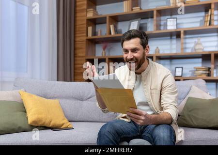 A joyful young man is sitting on the sofa at home and holding an envelope with a letter in his hands. Received good news, happy. Stock Photo