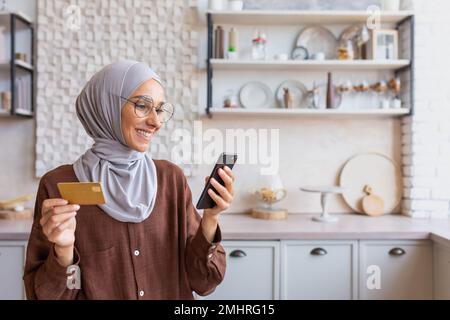 Young smiling Arab woman in hijab using phone and credit card at home in kitchen. Do online shopping. Stock Photo