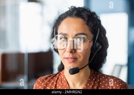 Close-up photo. Portrait of a young beautiful Latin American woman in a headset. Call center, service operator, hotline, support and assistance line. He looks seriously into the camera. Stock Photo