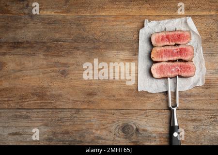 Carving fork with slices of delicious beef steak on wooden table, top view. Space for text Stock Photo