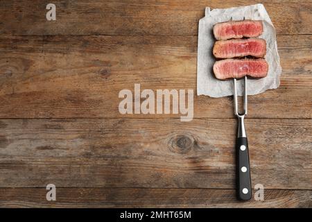 Carving fork with slices of delicious beef steak on wooden table, top view. Space for text Stock Photo