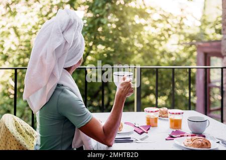 a middle-aged caucasian woman has breakfast on the terrace of her house after a shower. she is wearing a towel on her head. concept of well-being and Stock Photo