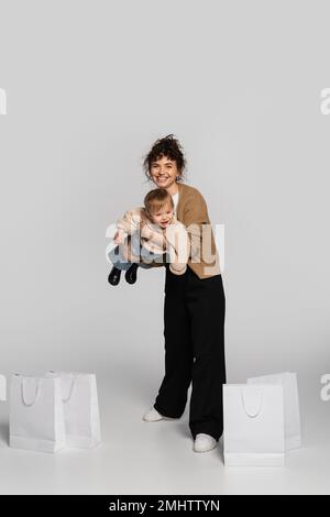 full length of cheerful woman in casual clothes holding in arms toddler girl near shopping bags on grey,stock image Stock Photo