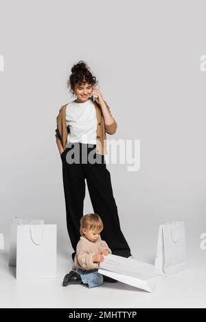 full length of cheerful woman talking on smartphone near toddler girl and shopping bags on grey,stock image Stock Photo