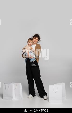 full length of cheerful mother in casual clothes holding in arms toddler girl near shopping bags on grey,stock image Stock Photo