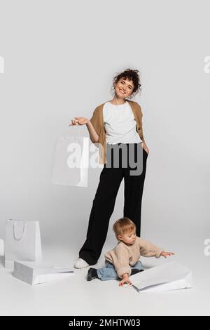 full length of cheerful woman in casual clothes posing with hand in pocket near toddler daughter and shopping bags on grey,stock image Stock Photo